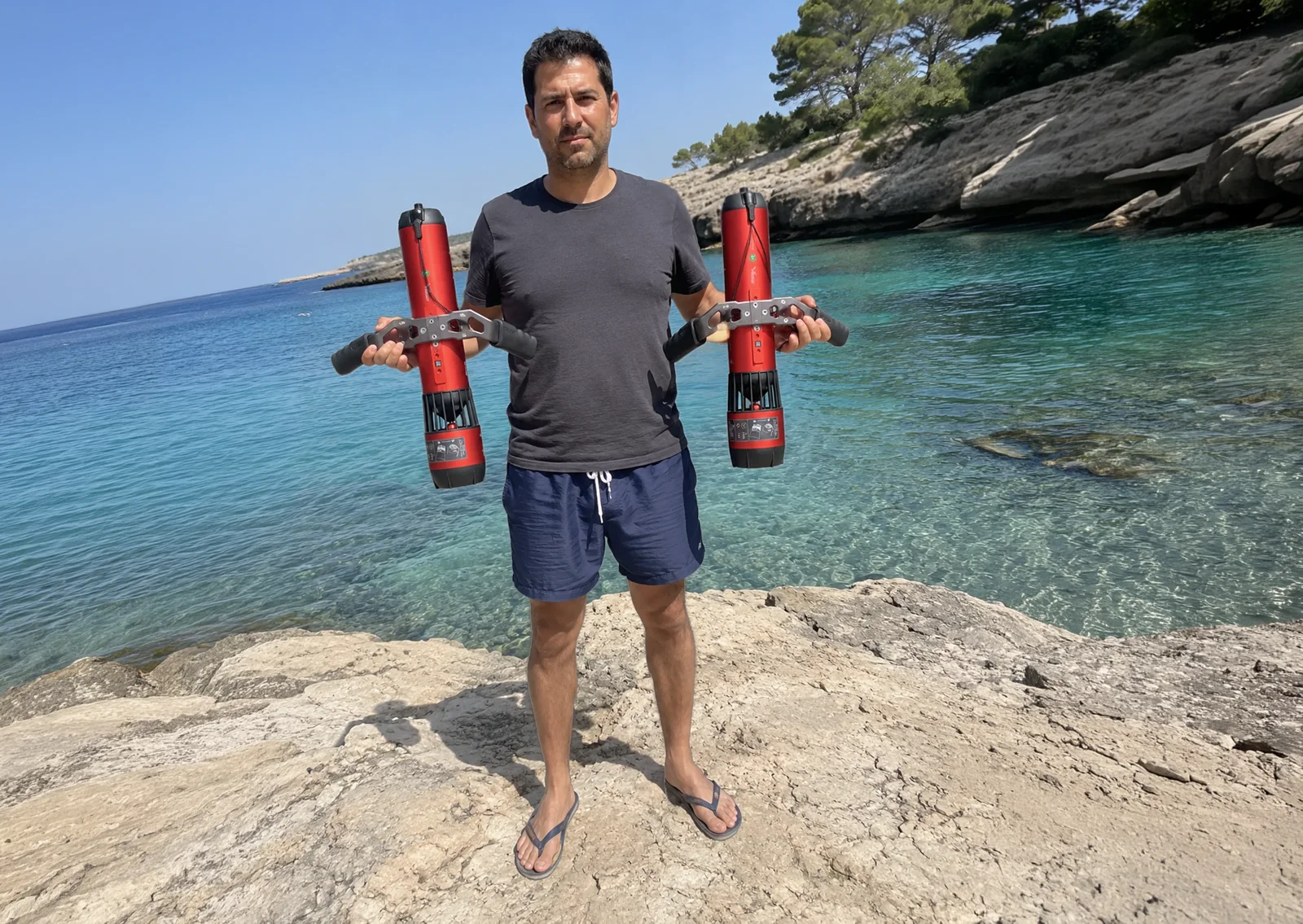 A man standing on the rocks of a turquoise cove holds two red underwater scooters, one in each hand.
